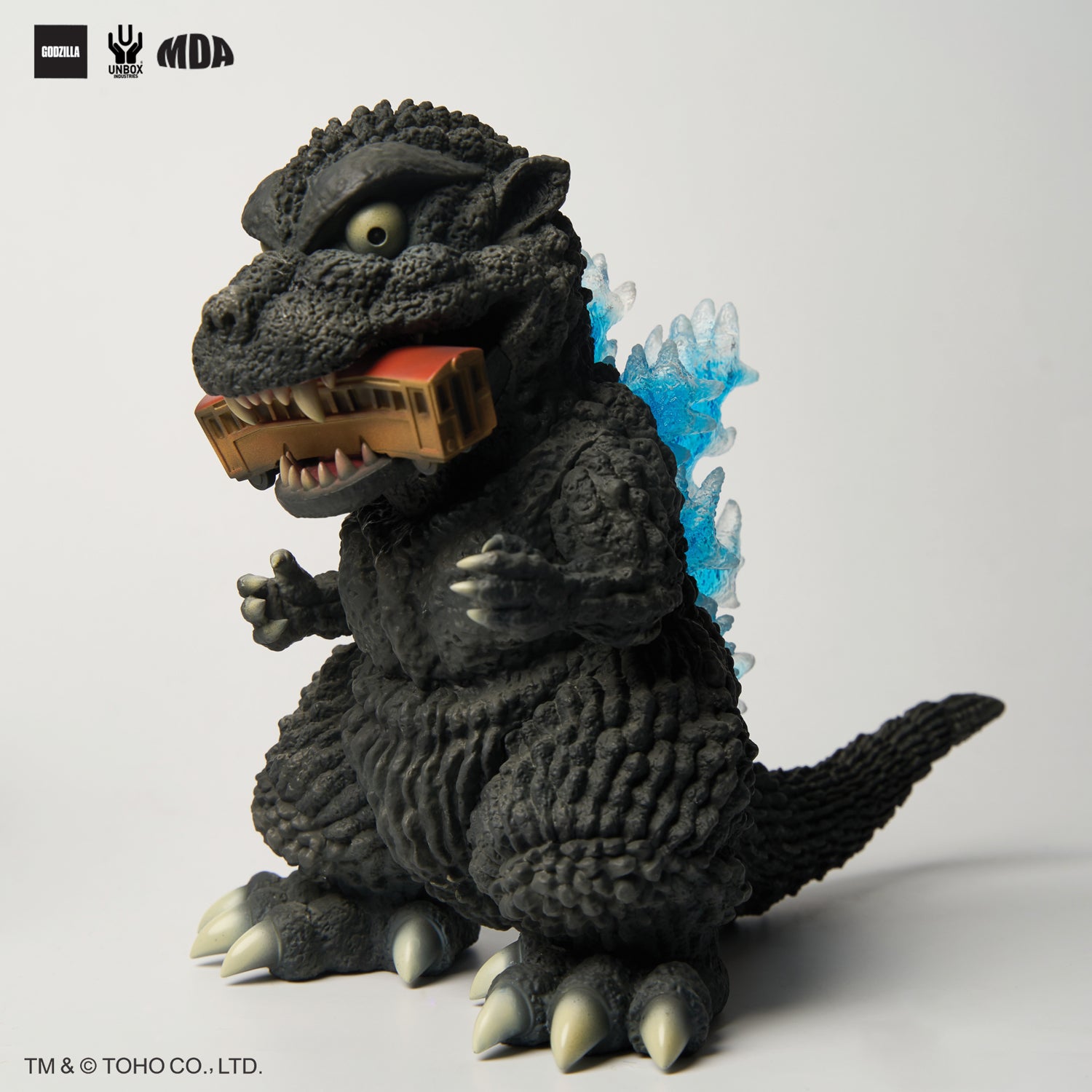 GODZILLA 1954 MDA PROJECT - Preorder: Stylized Godzilla vinyl figure with blue dorsal fins, gripping a train car, logos and copyright visible.