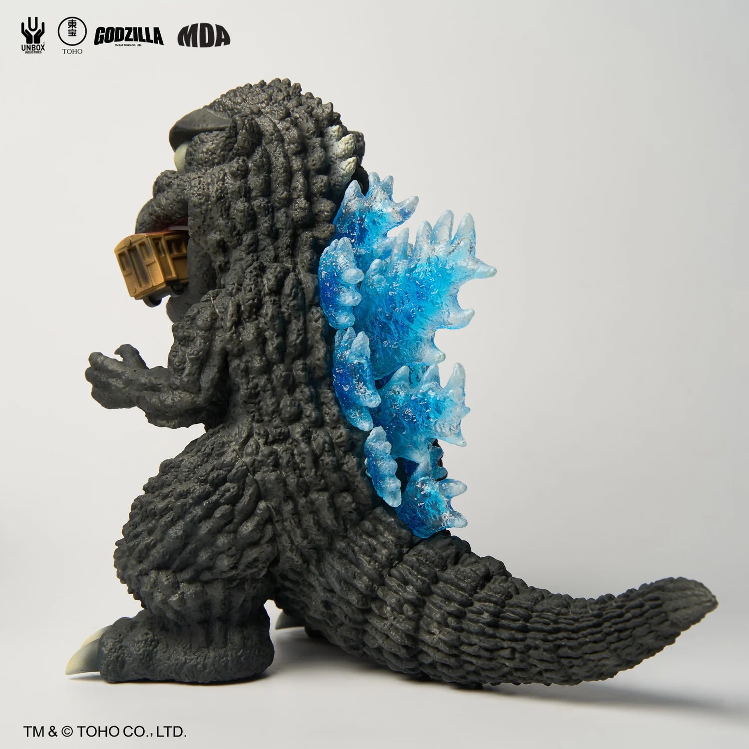 GODZILLA 1954 MDA PROJECT - Preorder: Slate-gray vinyl figure with textured skin and blue dorsal plates, holding a train carriage. Sculpted by MDA for Unbox Industries.