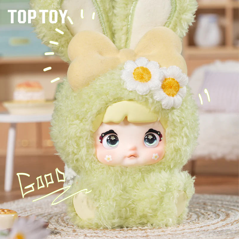 Nommi Baby Sweetheart Bunny Plush in a fluffy green suit, part of a blind box series with 8 designs, displayed on a woven mat.
