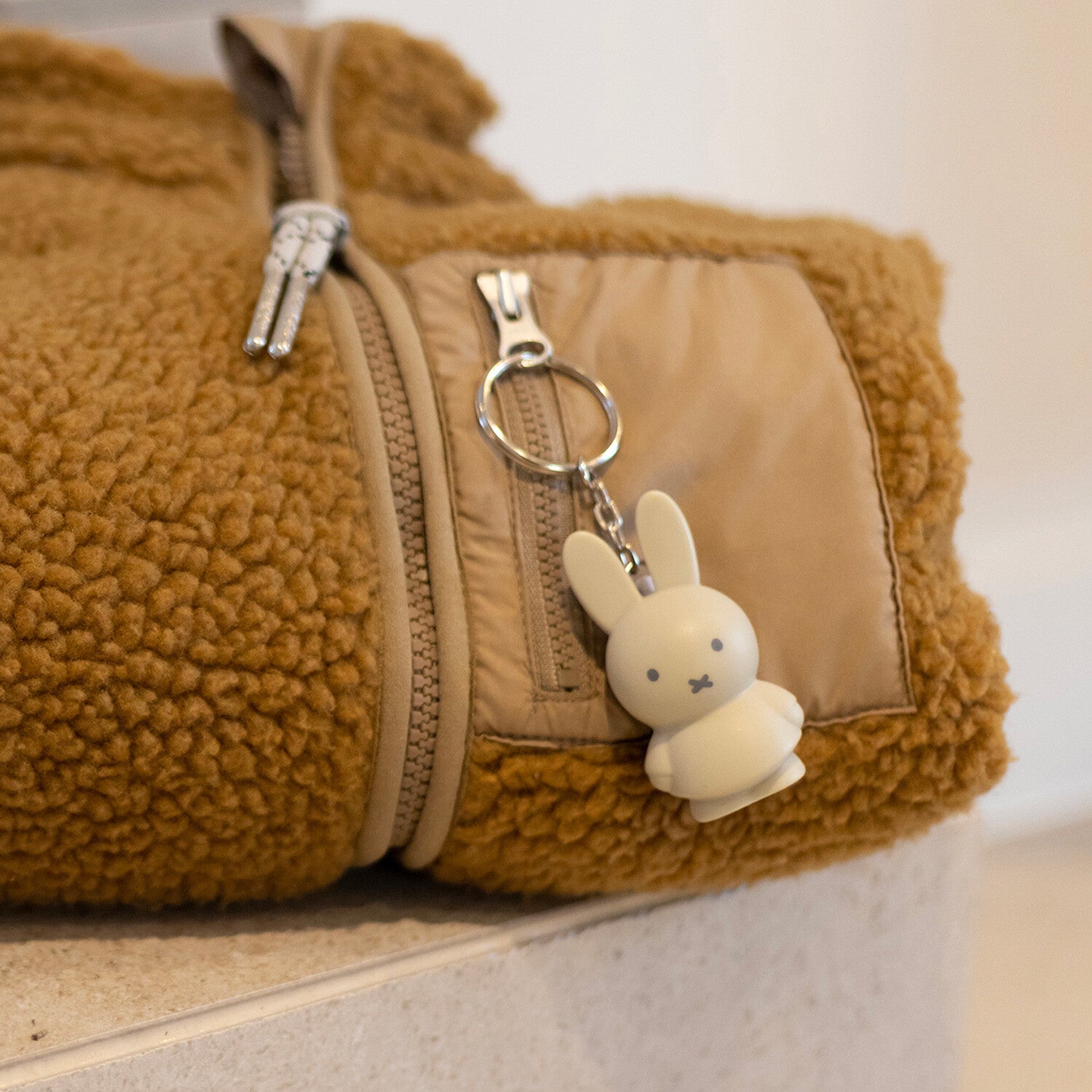 Miffy Keychain featuring a cute white bunny face, attached to a zipper on a blanket, suitable for ages 3 and up, CE certified.