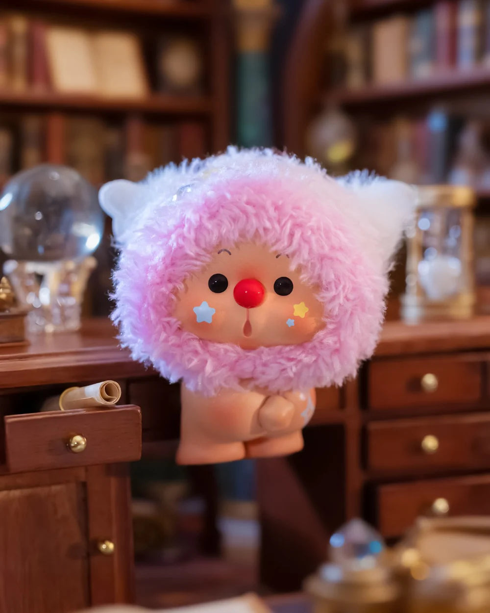 OYO Constellation Story Hipper Series figurine on desk, red-nosed with pink hood, surrounded by books, crystal ball, and hourglass, evoking a cozy wizard's study.
