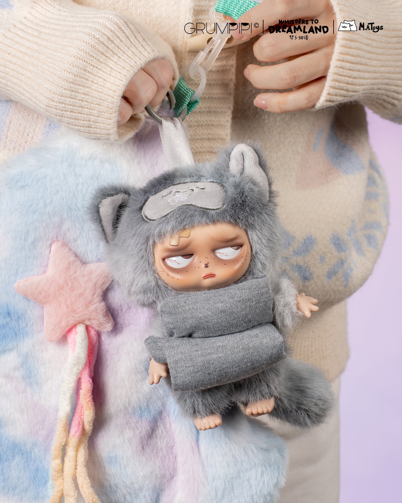 Hands hold a Sleepless Grumpipi Whispers to Dreamland plush keychain, featuring a fuzzy bag with a grumpy doll in a cat onesie and rainbow tassels.
