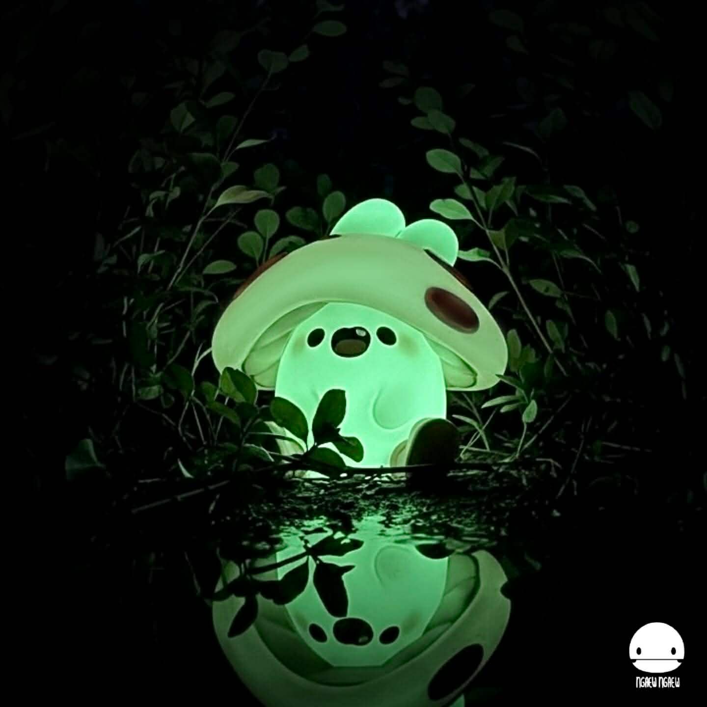 Ngaew Ngaew Poison Mushroom glow-in-the-dark resin lamp, shaped like a cartoon mushroom, emits soft green light, reflected in water, nestled among leaves.