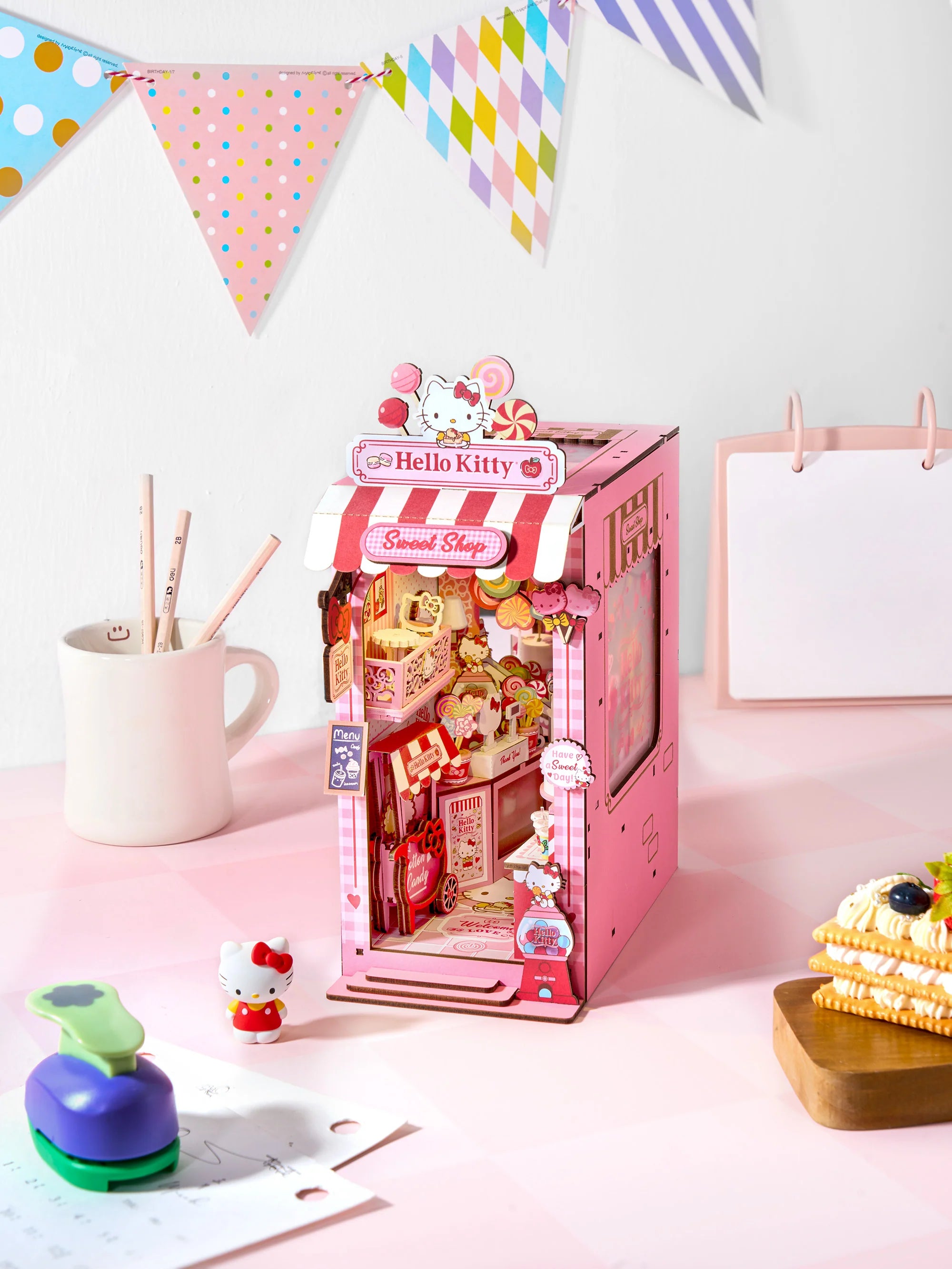 Hello Kitty® Sweet Shop Booknook diorama with candy props, Hello Kitty figurine, waffle cookies, and pencil mug on checkered tabletop.