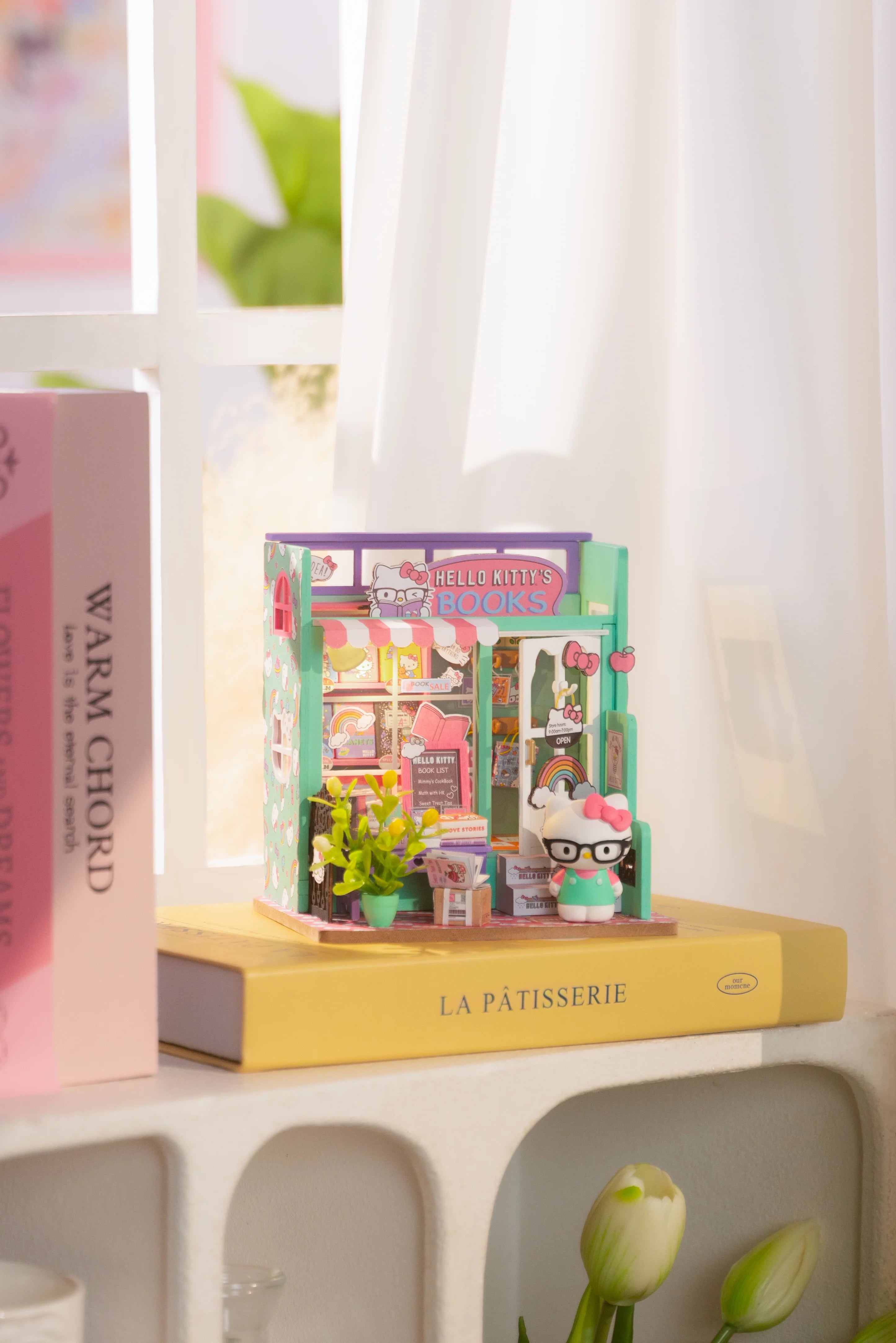 Hello Kitty® Bookstore diorama atop real books, featuring tiny books, potted plants, and a Hello Kitty figure with glasses, embodying whimsical craft charm.