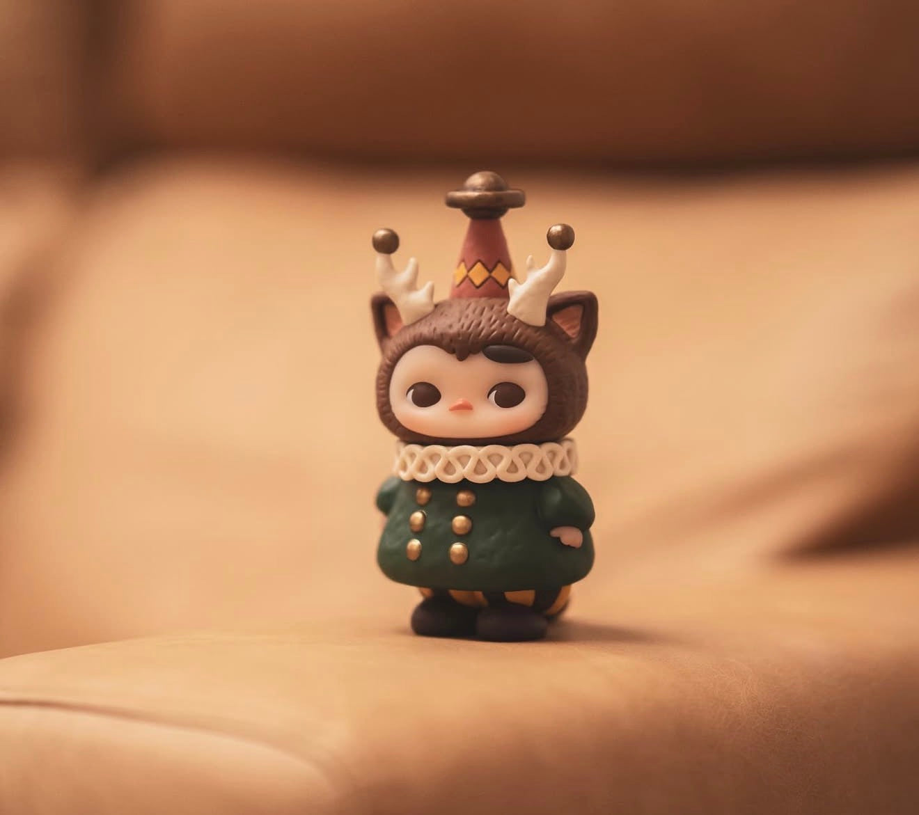 Pucky x Kaori Morris Pucky vinyl figurine on a couch cushion, featuring a green coat, white collar, and animal hood with antlers and a party hat. Preorder available.