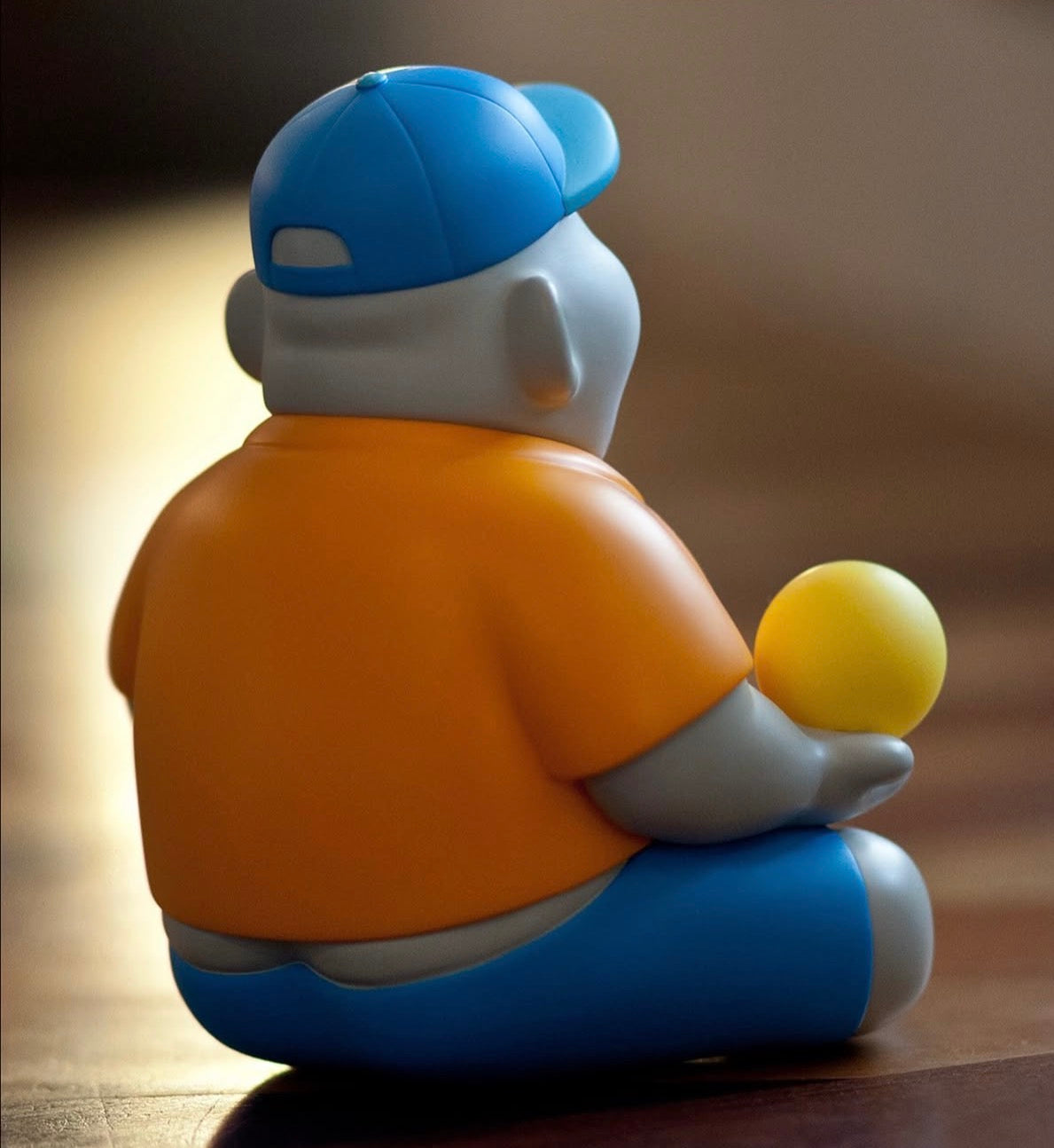 Miller by Jim Dreams figure, 13.5cm, seated with a yellow ball, wearing an orange T-shirt and blue cap, on a wooden floor.