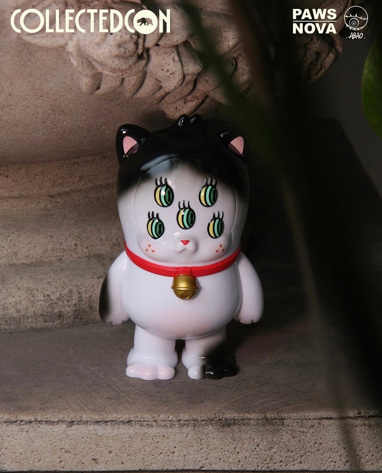 CollectedCon Abao Beans Exclusive vinyl toy, a six-eyed cat with black paint and a red collar, on stone steps. Preorder for May 2026 release.