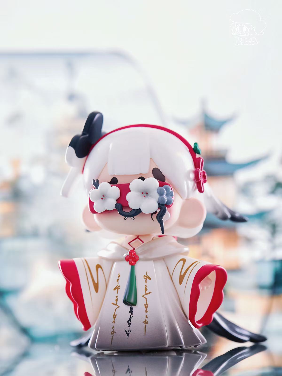 KASA 2.0 - Mystic Tales figure in a white and red kimono with gold script, featuring a green pendant, floral headband, and eye blossoms, standing on a reflective surface.