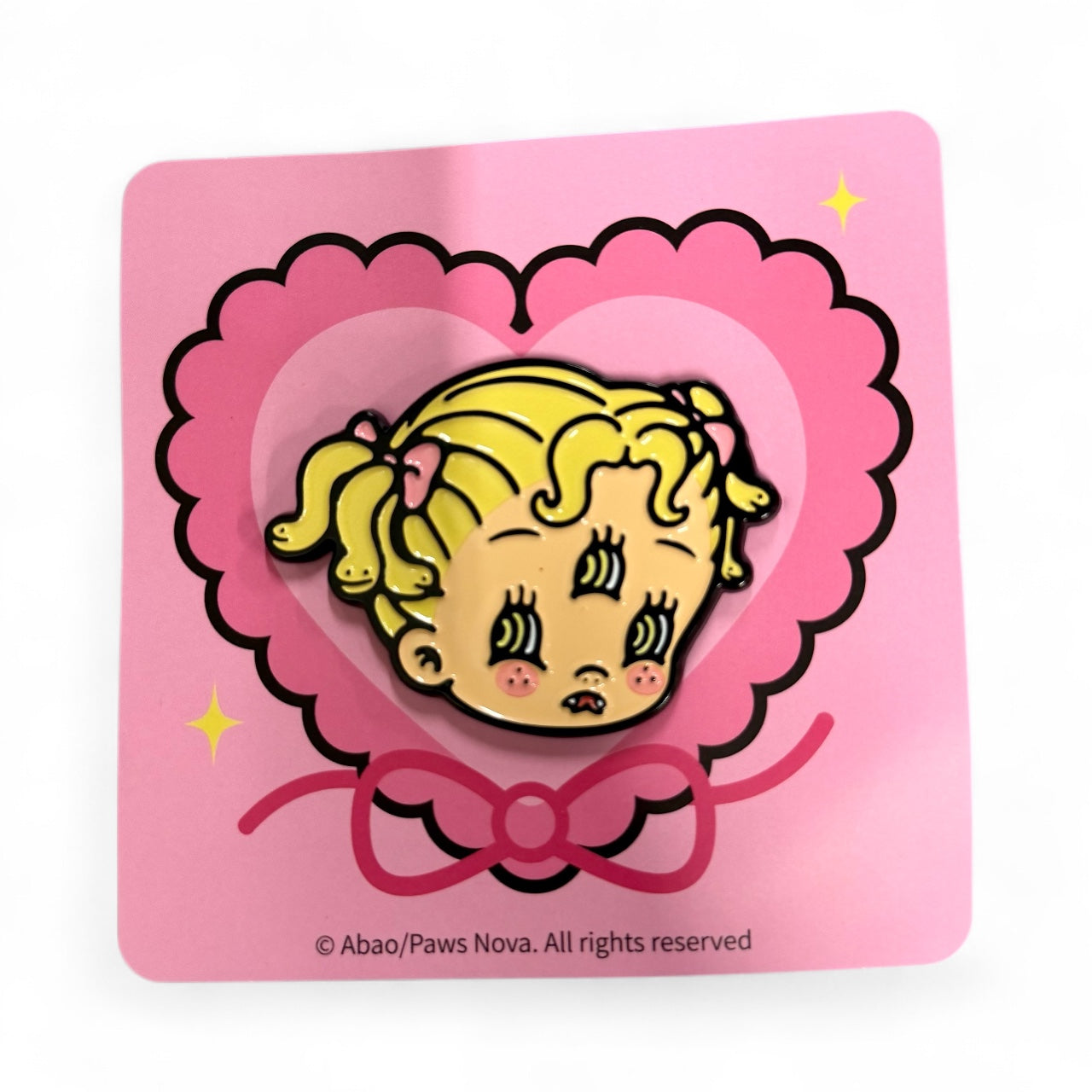 Baby Dassi Metal Badge: An enamel pin featuring a surprised blonde girl with pigtails, mounted on a pink heart-themed card, from Strangecat Toys.