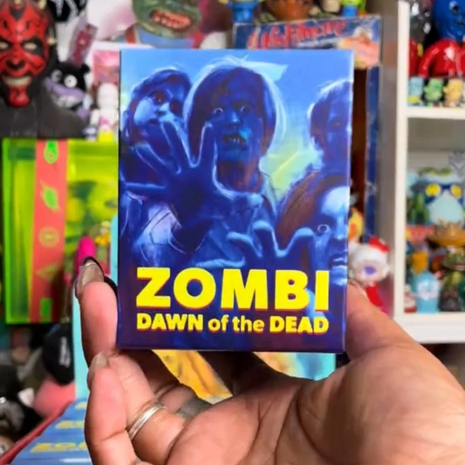 Person holding ZOMBI: DAWN OF THE DEAD Who Goes There Blind Box Series, featuring zombie artwork; shelves with toys visible in background.