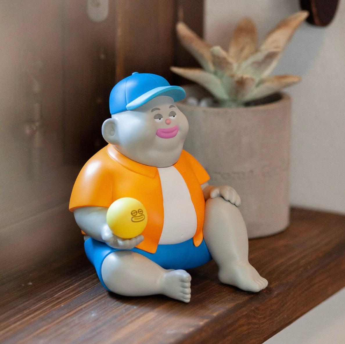 Miller by Jim Dreams vinyl figurine, 13.5cm, with gray skin, wearing an orange shirt, blue shorts, and cap, holding a yellow ball, sitting on a shelf.