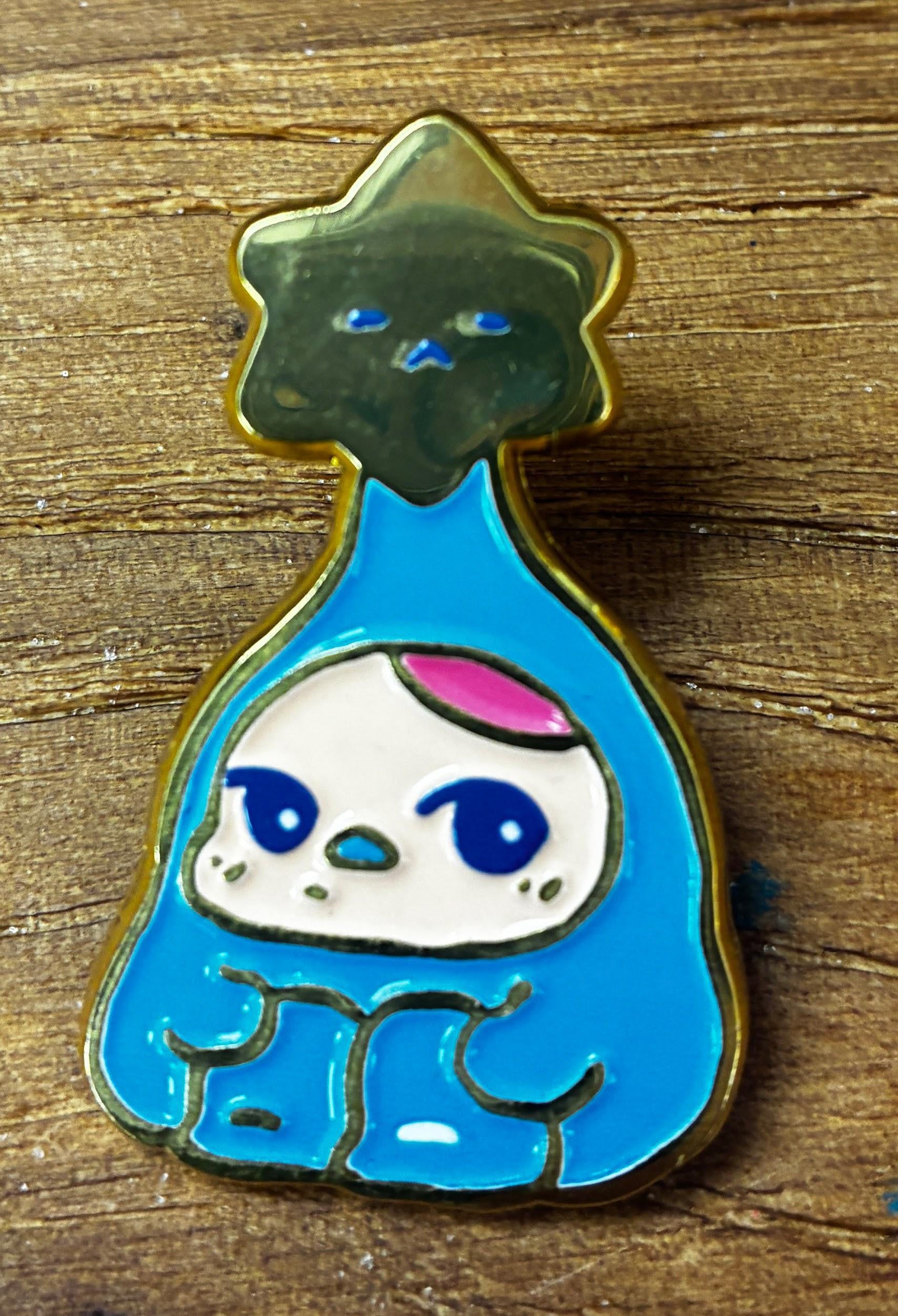 Bloom Pins by Pucky: A stylized creature in a blue cloak with a gold star, featuring big eyes and a tiny beak-like nose.