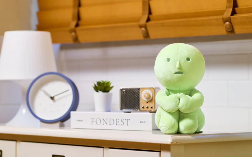 SMISKI Plush sits on a book beside a tabletop radio, succulent, lamp, and clock, offering comfort and charm in a Strangecat Toys setting.
