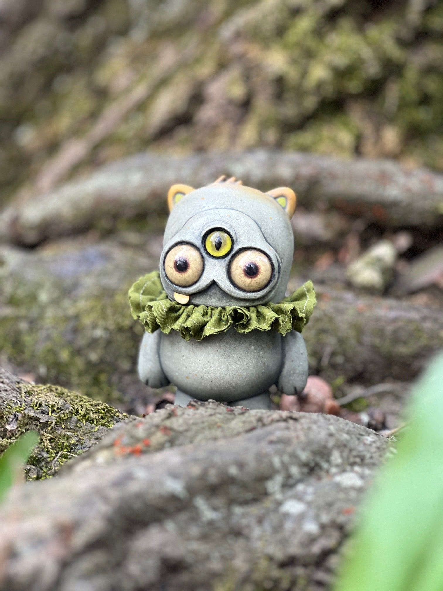 Ruffletoof Beans figure by Chris Ryniak x Amanda Louise Spayd x Josh Divine, a three-eyed, cat-like vinyl toy, on mossy stone. Preorder for May 2026.