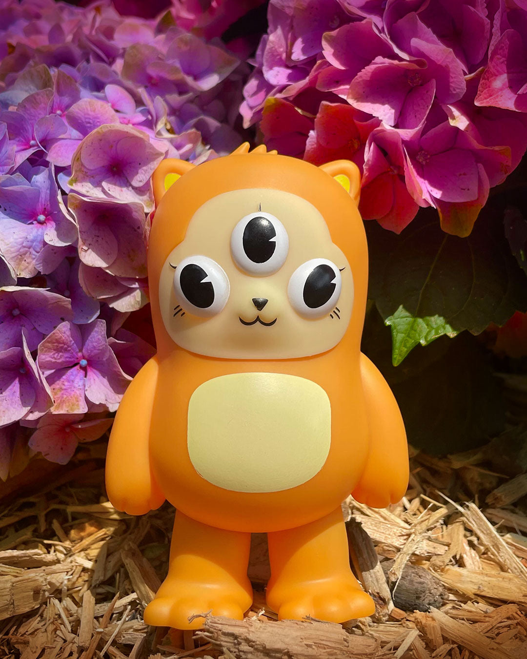 Beans - I Hate Mondays Exclusive: A 5 orange vinyl toy with three eyes and a yellow belly, standing on mulch in front of hydrangeas.